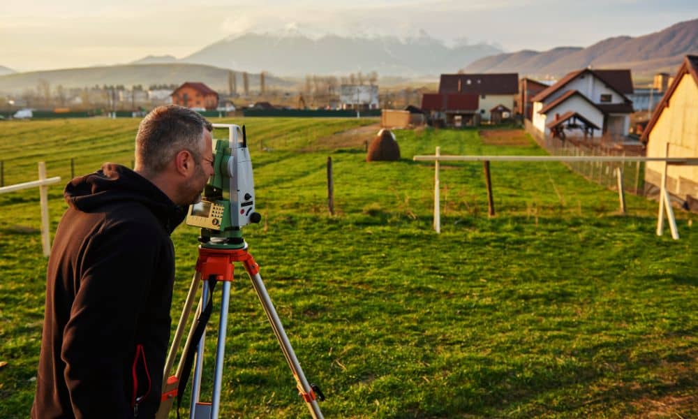 Licensed land surveyor measuring property boundaries in a residential area with mountains in the background.