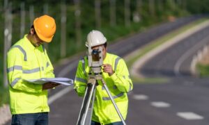 Survey crew performing land surveying near a roadway while wearing high-visibility safety gear